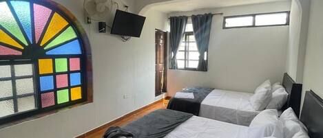 A hotel room with two beds, a large window with colorful stained glass, a ceiling fan, and a flat-screen TV mounted on the wall.
