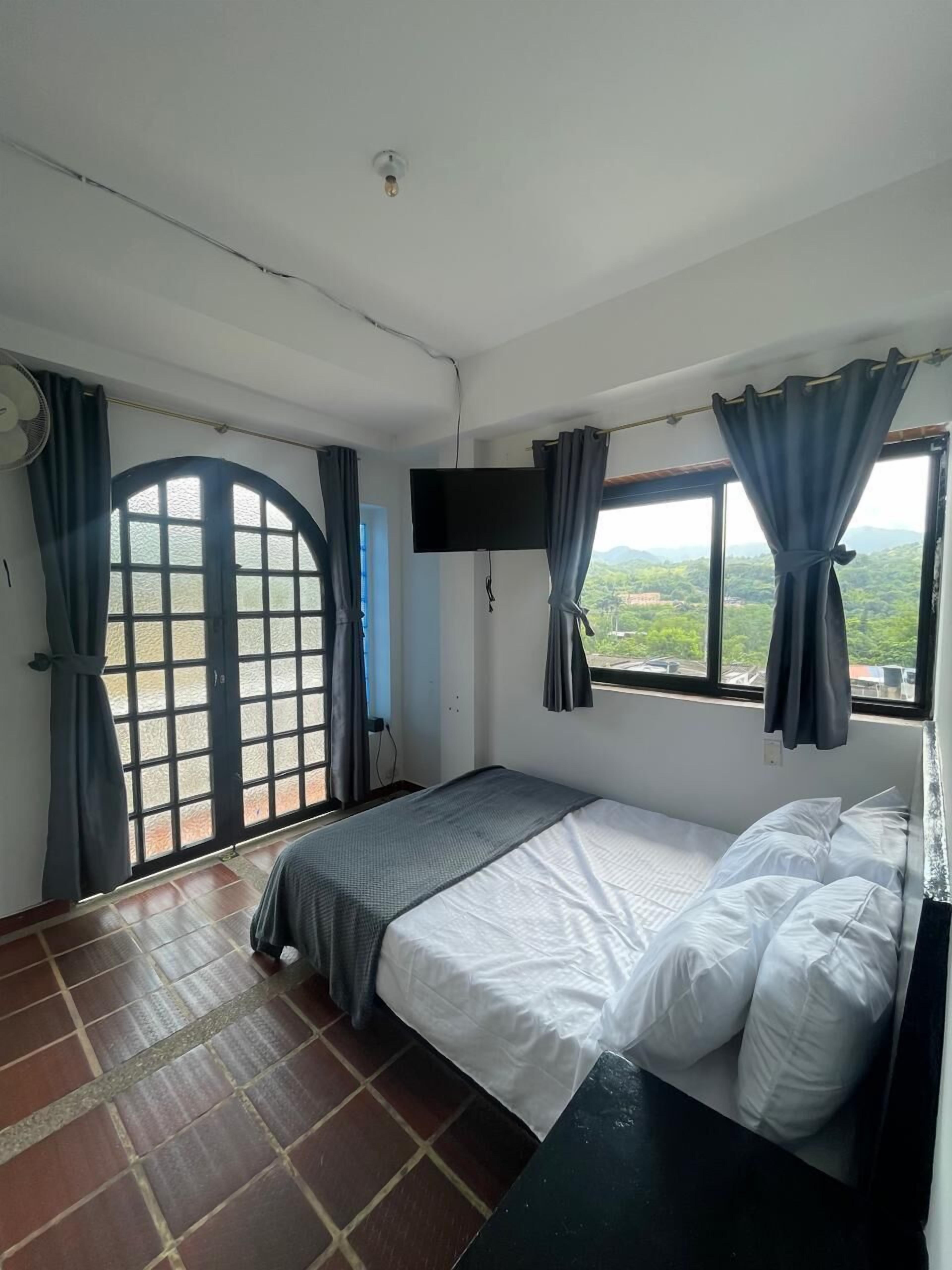 A bedroom with a bed, a TV, a window with curtains, and a view of greenery outside.