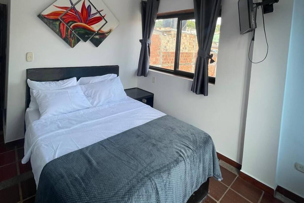 Une chambre à coucher avec un lit, un téléviseur fixé au mur, une fenêtre avec des rideaux et un tableau accroché au mur.