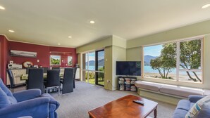 A living room with a sofa, a coffee table, and a TV. There is a dining area with chairs and a table. A bookshelf is visible, and a window offers a view of trees and a beach.