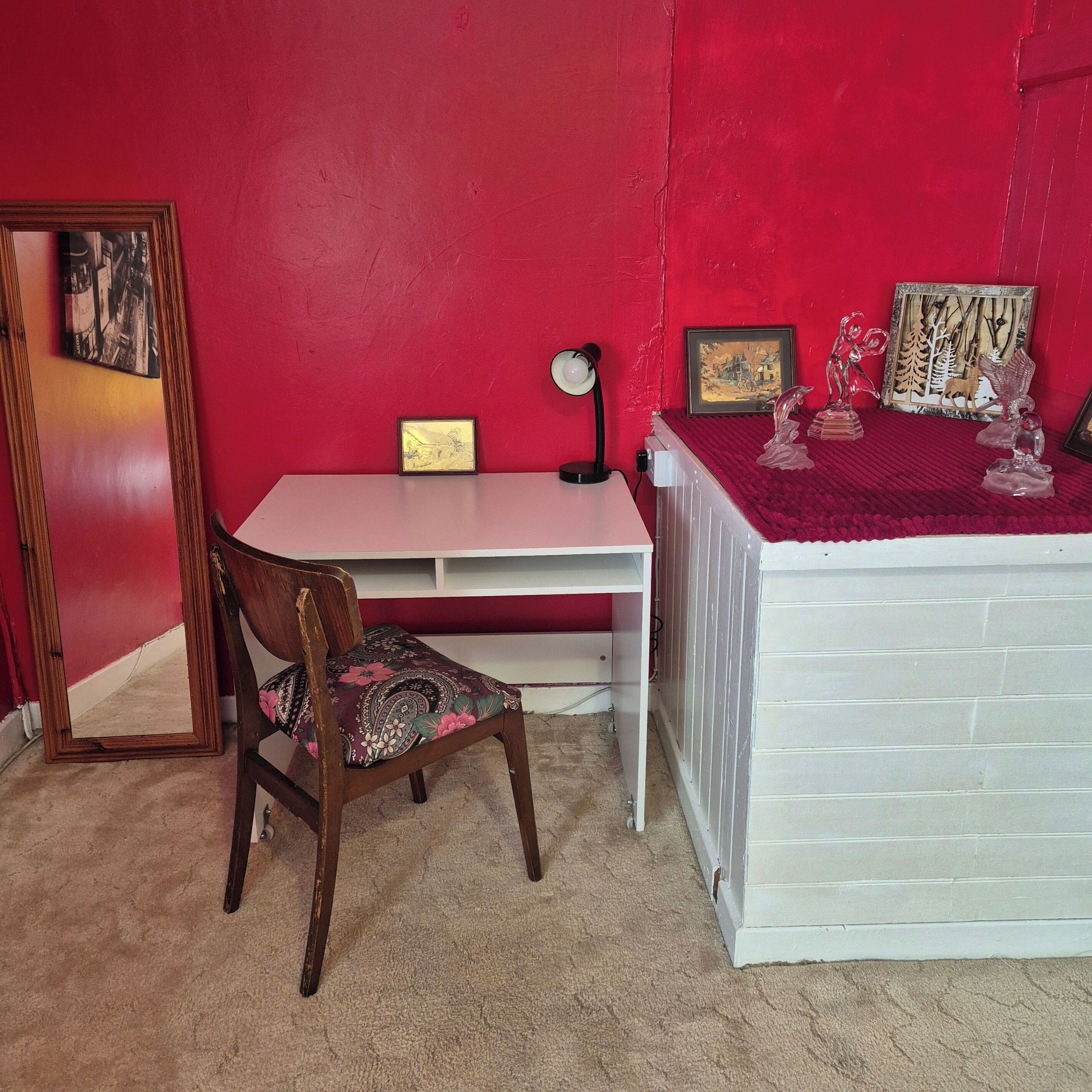 A room with a red wall, a white desk, a wooden chair, and a white bed.