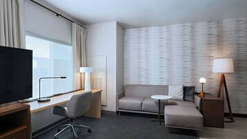Una habitación de hotel moderna con un televisor de pantalla plana grande, un escritorio con lámpara, una silla de oficina gris, un sofá, una mesita redonda y dos lámparas de pie.