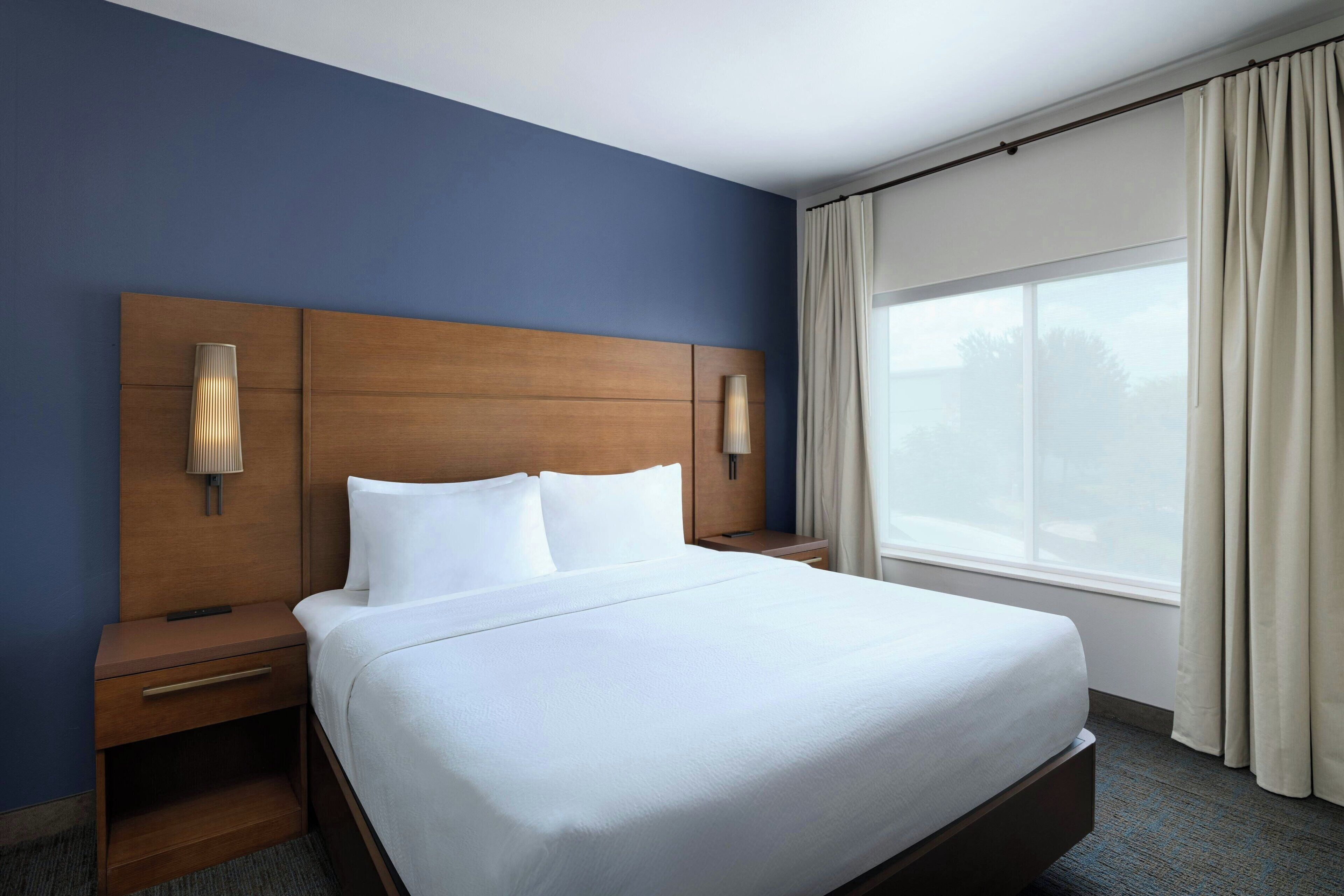 A hotel room with a large bed, wooden headboard, two bedside tables with lamps, a window with curtains, and a view of the outside.