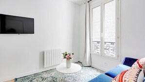 Una sala de estar moderna con un televisor de pantalla plana montado en la pared, una mesa de centro redonda blanca con un florero, un sofá azul con cojines estampados y un ventanal con vistas al balcón.