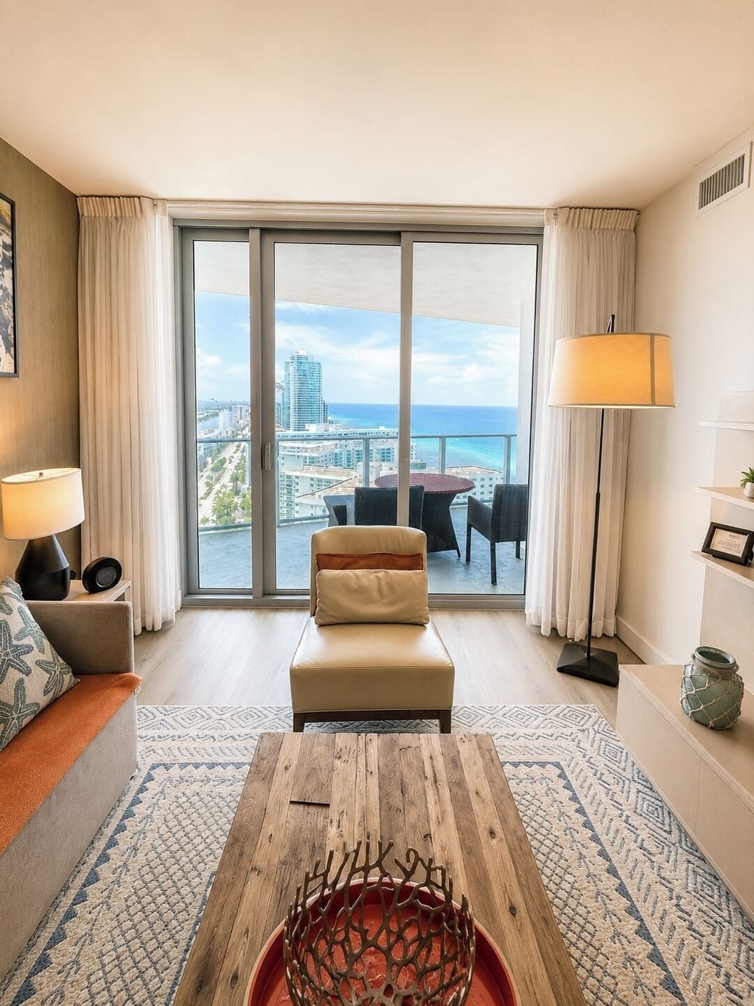 A hotel room with a large sliding glass door to the balcony, a sofa, a coffee table, and a lamp.
