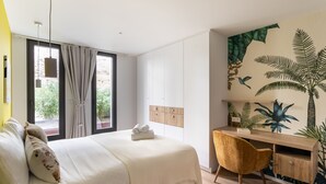 A bedroom with a bed, a desk, and a chair. There is a window with curtains, a wall with a tropical mural, and a wooden headboard. - La Briouette - avec vue panoramique d’exception (Pléneuf-Val-André)