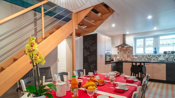 A dining area with a red tablecloth, white chairs, and a potted orchid. A staircase with wooden railings is visible in the background. - Hon Ty Glas - Maison proche de plage avec jardin (Moëlan-sur-Mer)