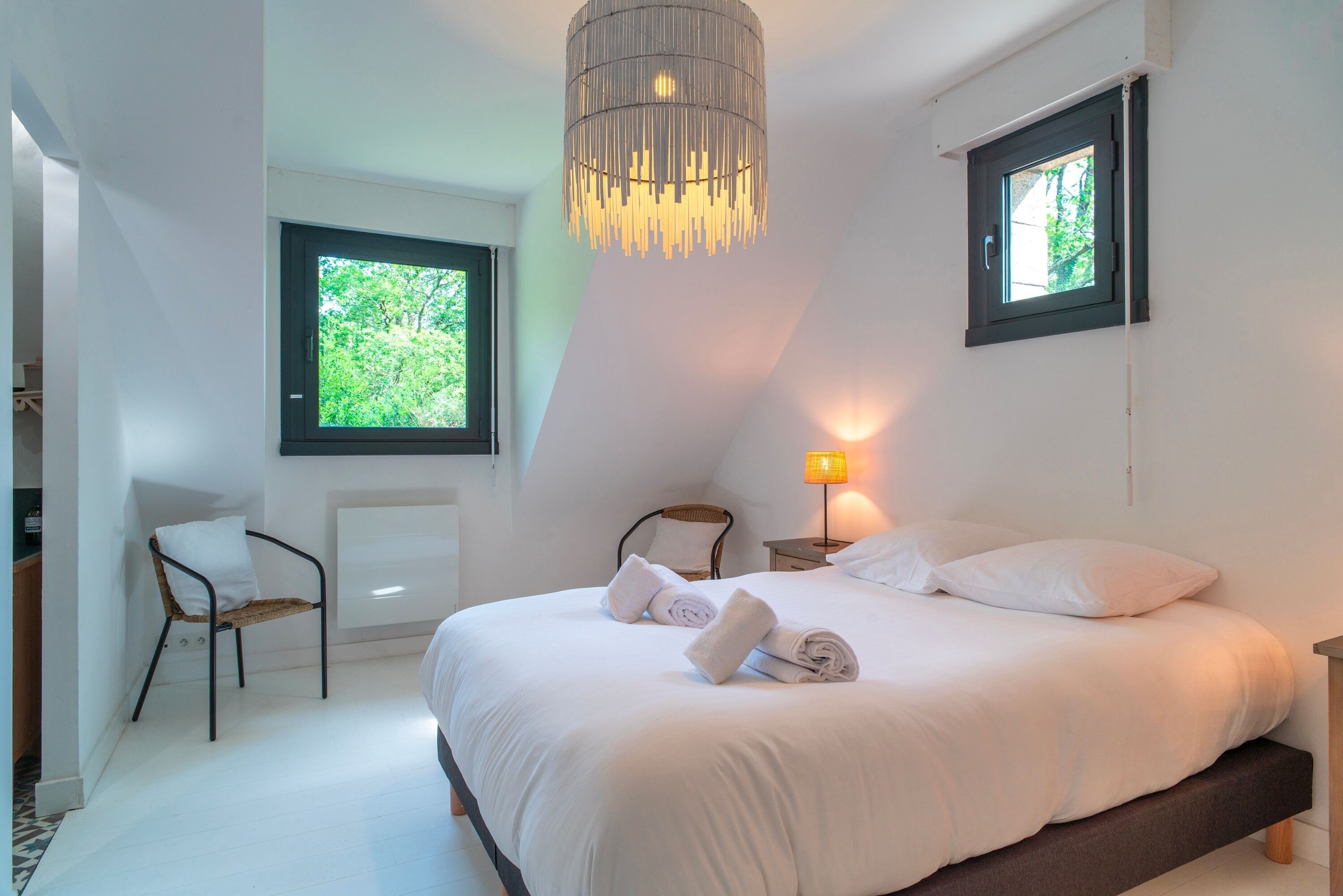 A modern bedroom with a large bed, a hanging light fixture, a window with a view of greenery, and a chair.