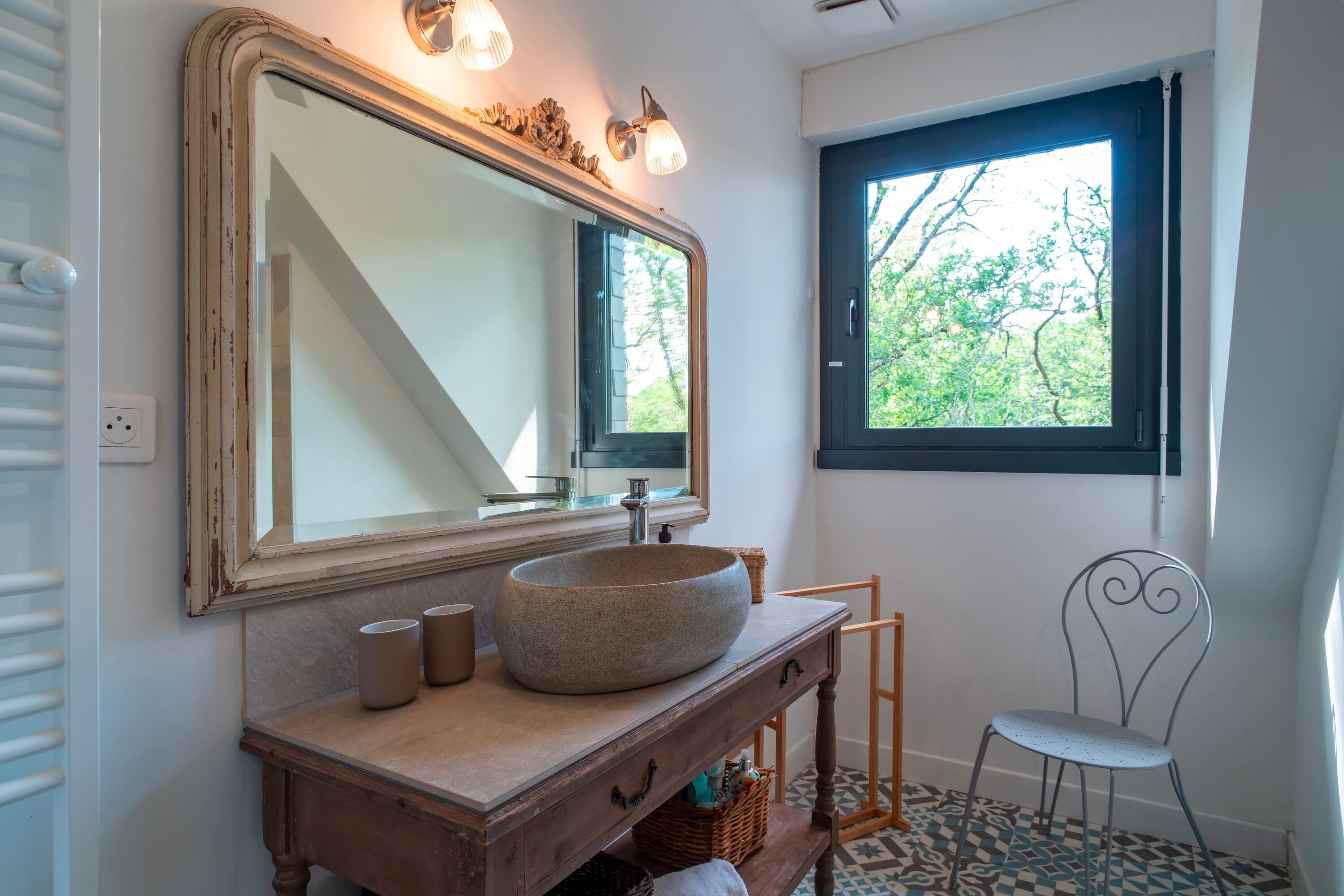 A bathroom with a large mirror, a round sink, and a window with a view of trees.