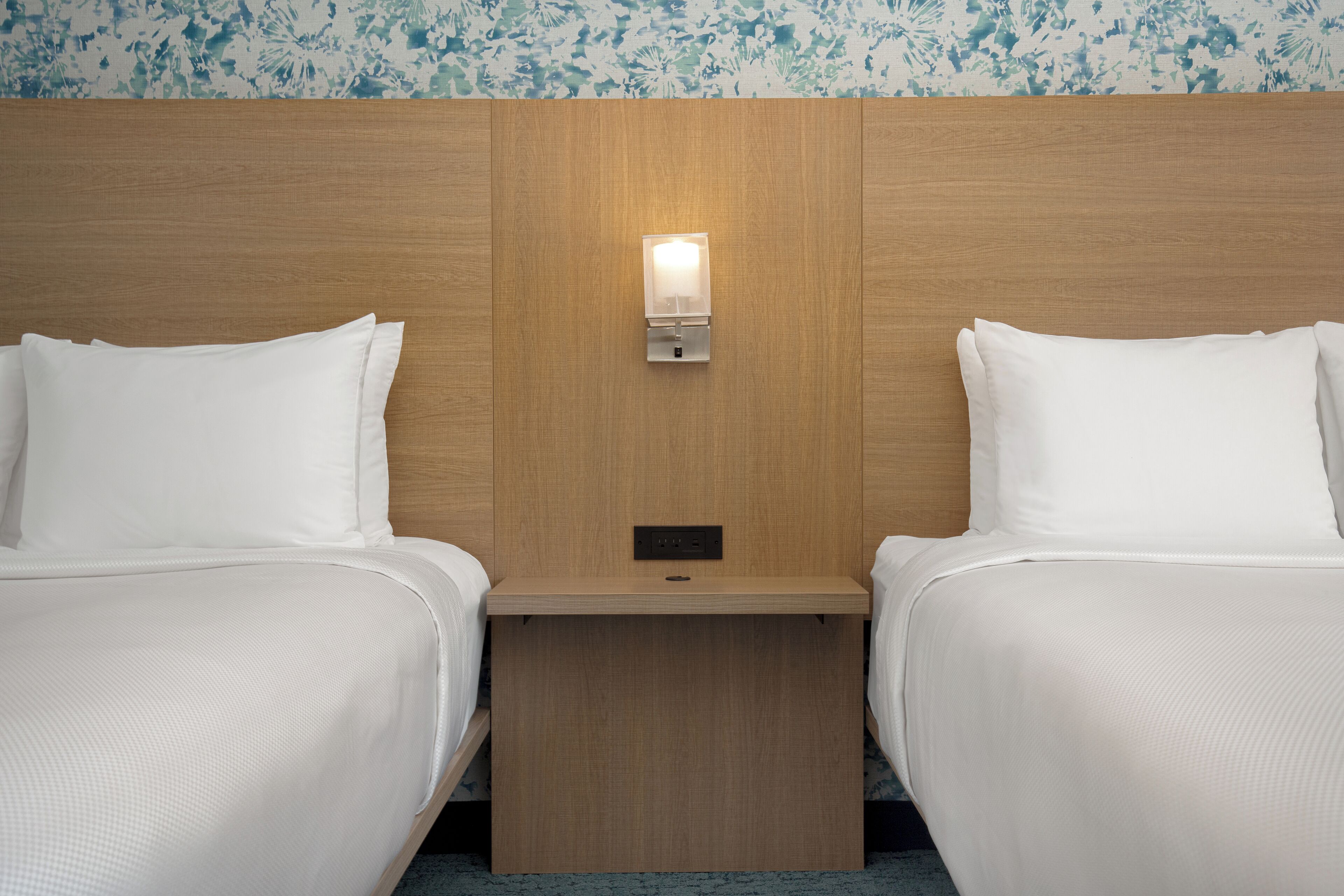 Una habitación de hotel con dos camas, un cabecero de madera, una mesita de noche con lámpara y un papel tapiz estampado con motivos florales.
