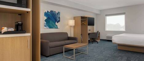 A hotel room with a sofa, a small table, a desk with a chair, a bed, a wardrobe, a coffee maker, and a painting on the wall.