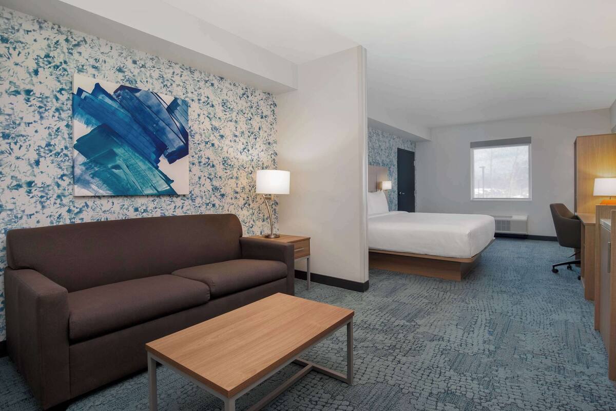 A hotel room with a brown sofa, a wooden table, a bed with a white mattress, a desk with a chair, and a large abstract painting on the wall.