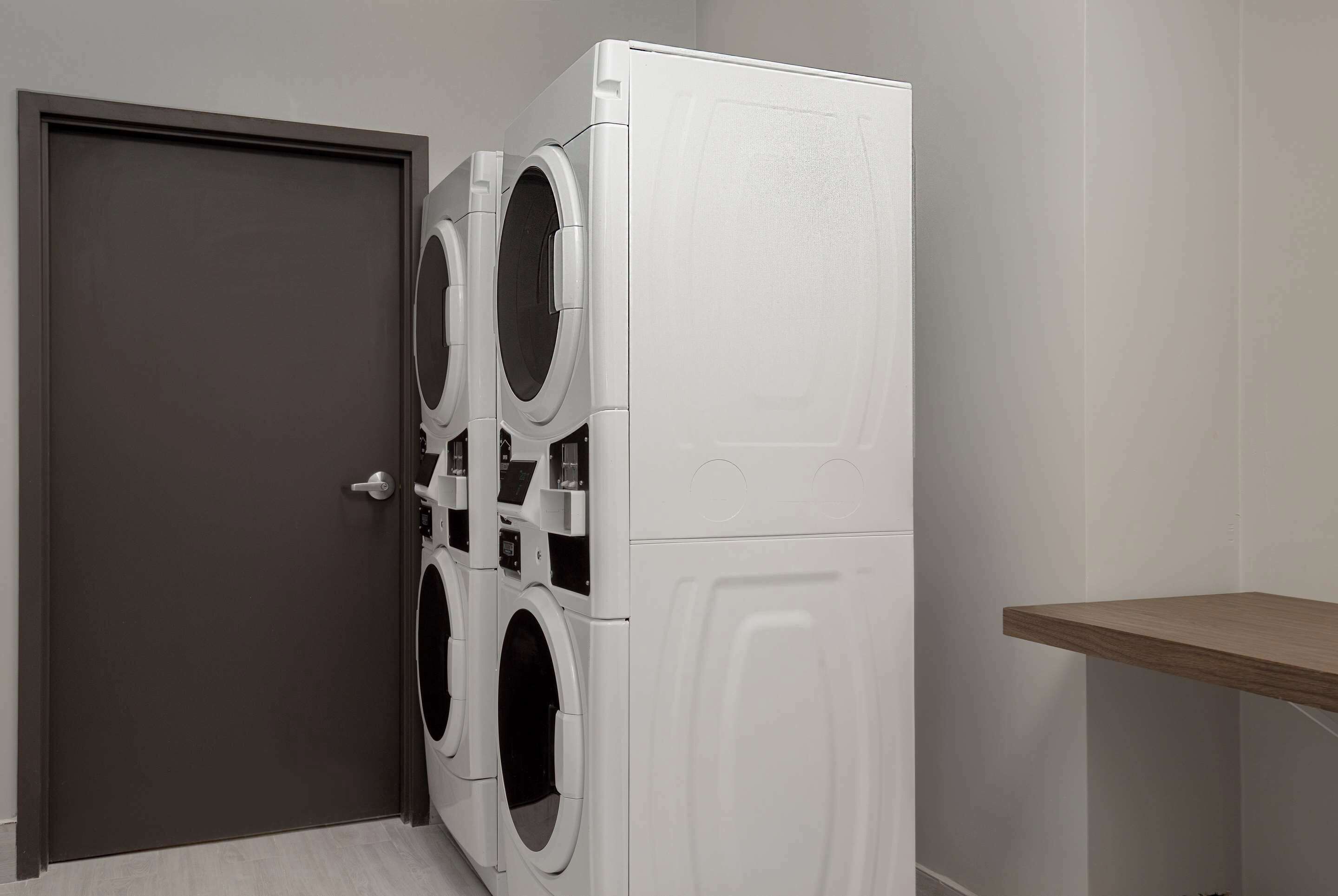 A laundry room with a door, a stacked washer and dryer unit, and a wooden shelf.