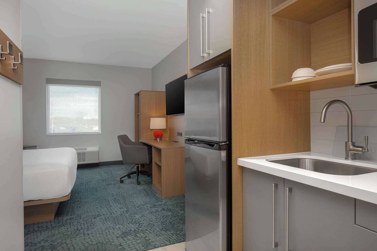 A compact hotel room with a bed, a desk with a chair, a refrigerator, a sink, and a window with a view.