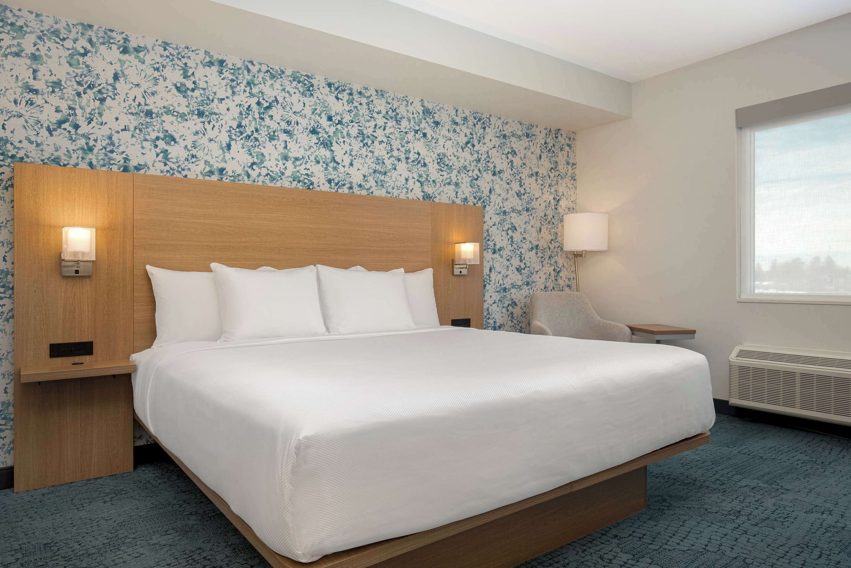 A hotel room with a large bed, two bedside tables, a nightstand, a wall-mounted lamp, and a window with a view.