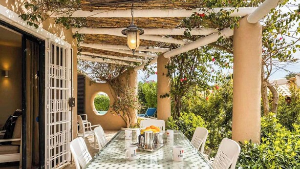 Un tavolo in vetro con sedie bianche sotto una pergola, immerso nel verde e con una piscina.
