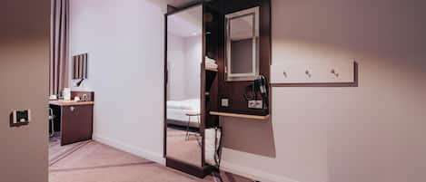 A hotel room with a bed, a bedside table, a mirror, a coat rack, and a small shelf.