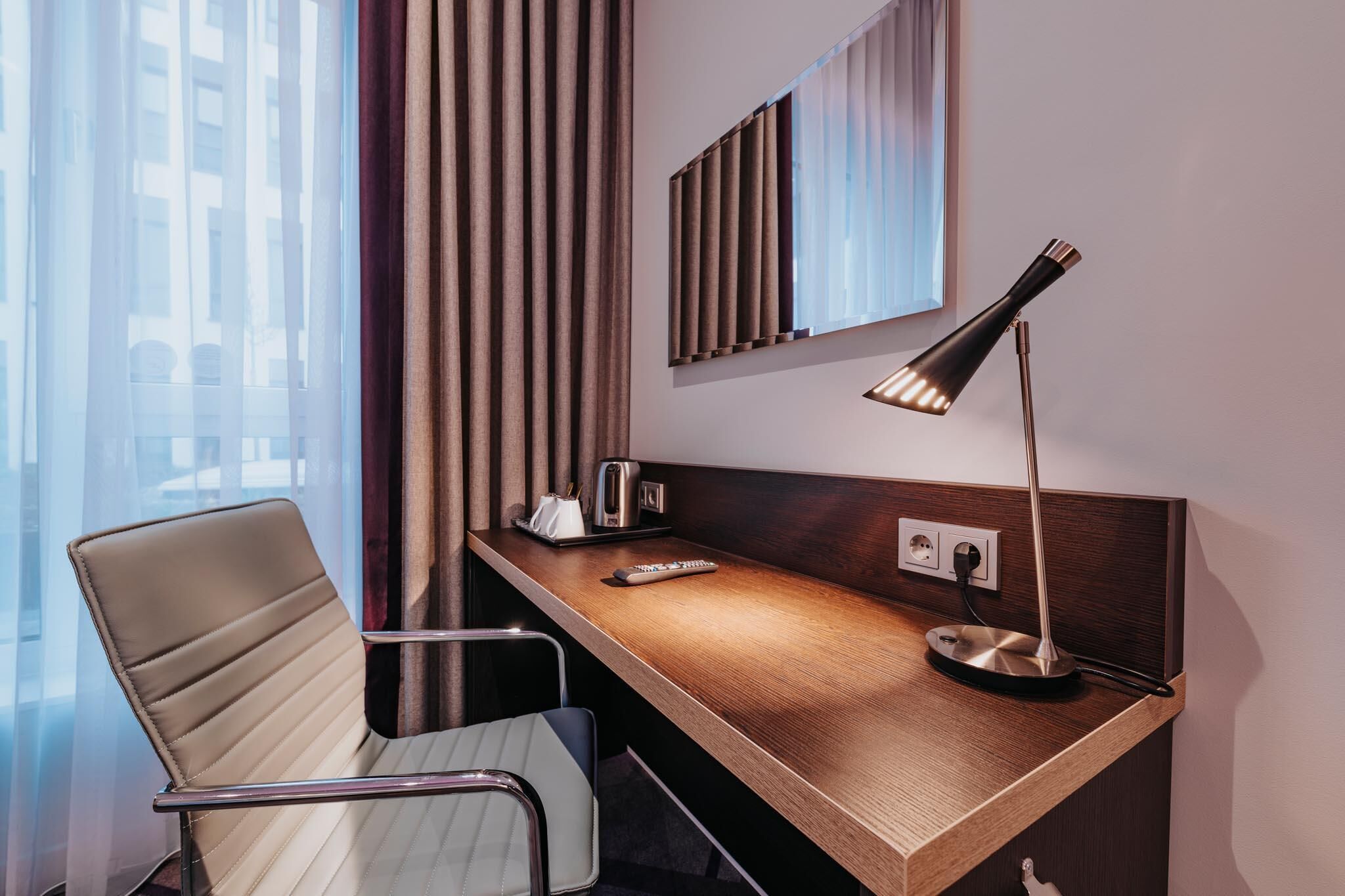 A modern hotel room with a wooden desk, a chair, a lamp, a mirror, and a window with curtains.