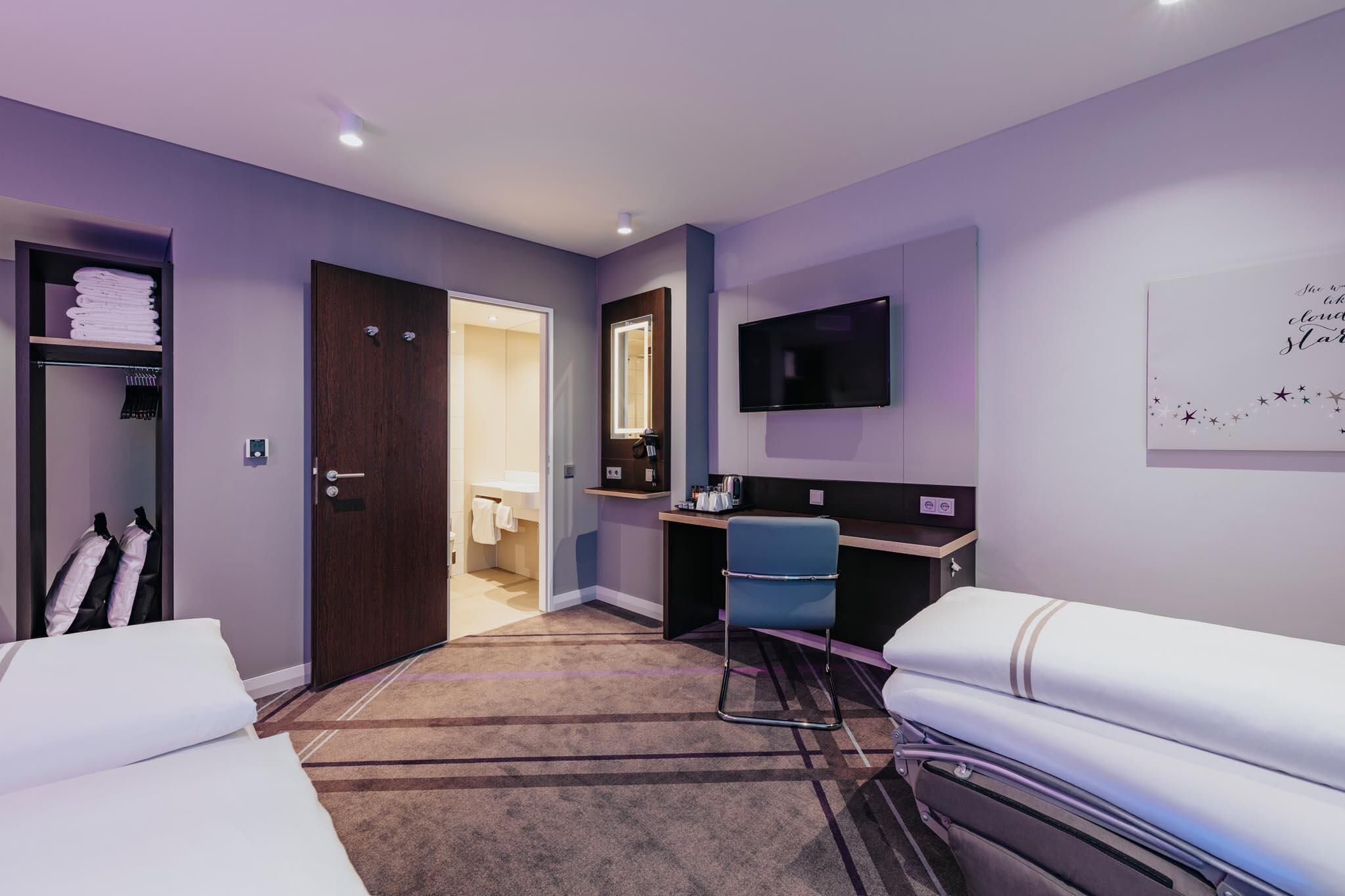 Una habitación de hotel con una cama, un escritorio con silla, televisión, espejo y armario.
