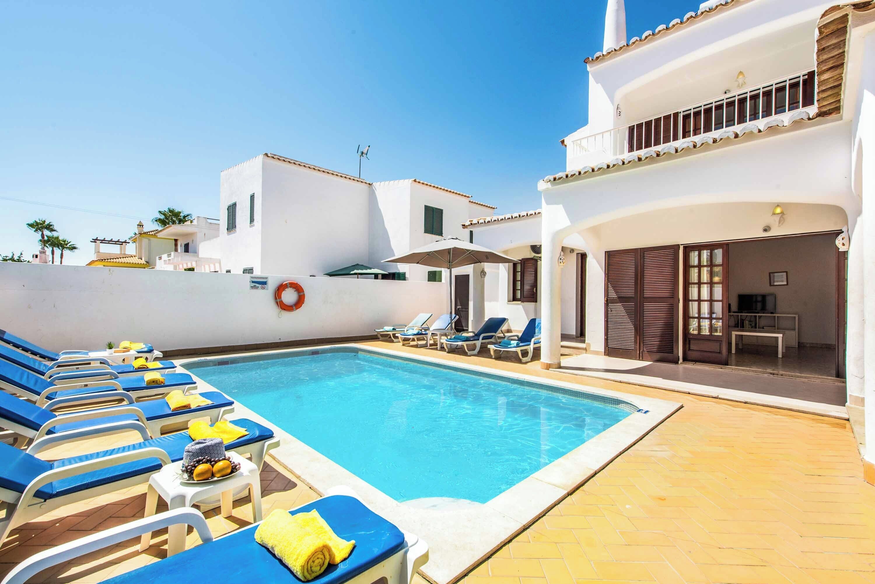 A swimming pool with sun loungers and umbrellas, a lifebuoy, and a building with balconies.