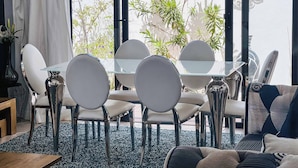 Un comedor con una mesa de vidrio y sillas blancas, un sofá con cojines estampados, y vista a zonas verdes exteriores.