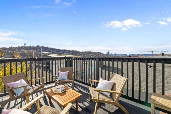 A balcony with wicker chairs and a table, overlooking a cityscape with buildings and a distant hill.