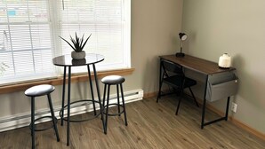 Une petite pièce bien éclairée, avec un bureau en bois, deux tabourets, une plante en pot et une lampe.