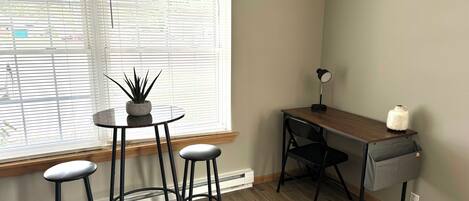 Una habitación pequeña y bien iluminada con un escritorio de madera, dos taburetes, una planta en maceta y una lámpara.