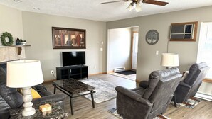 Un salon avec un ventilateur de plafond, un téléviseur, des fauteuils en cuir, une table basse en marbre et un tapis.