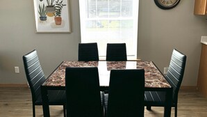 Un coin repas comprenant une table en marbre, quatre chaises noires, une œuvre d’art représentant une plante encadrée, une fenêtre avec des stores et une horloge murale.