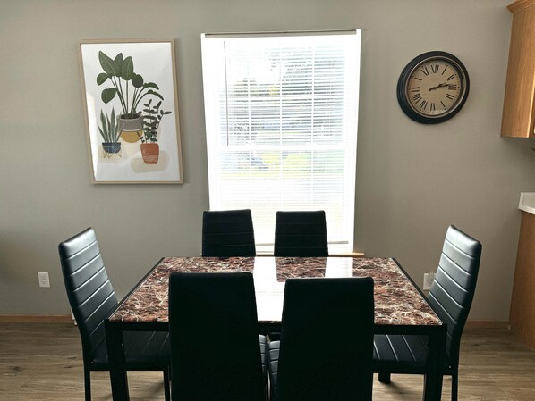 Een eetgedeelte met een marmeren tafel, vier zwarte stoelen, een kunstwerk van een plant in een lijst, een raam met zonwering en een wandklok.