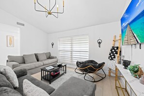 Un salon moderne comprenant un canapé, une table basse, un fauteuil à bascule et un téléviseur diffusant une scène tropicale.