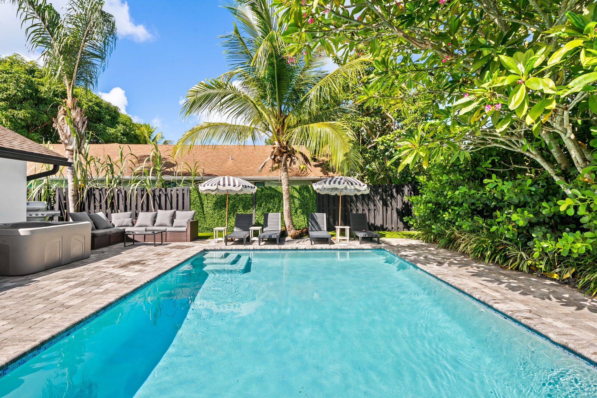 A rectangular swimming pool with a tiled edge, surrounded by lounge chairs and umbrellas, with a wooden fence and greenery in the background.