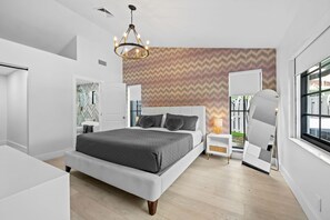 A modern bedroom with a large bed, a chandelier, a patterned wall, and a window with a view. - Casita Delray Oasis: 4BR with Pool, Hot Tub, BBQ, Firepit, Pool Table (Delray Beach)