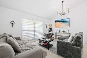 A modern living room with a sofa, armchair, coffee table, and a TV displaying a beach scene. - Casita Delray Oasis: 4BR with Pool, Hot Tub, BBQ, Firepit, Pool Table (Delray Beach)