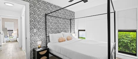 A modern bedroom with a four-poster bed, a ceiling fan, and a view of greenery through a window.