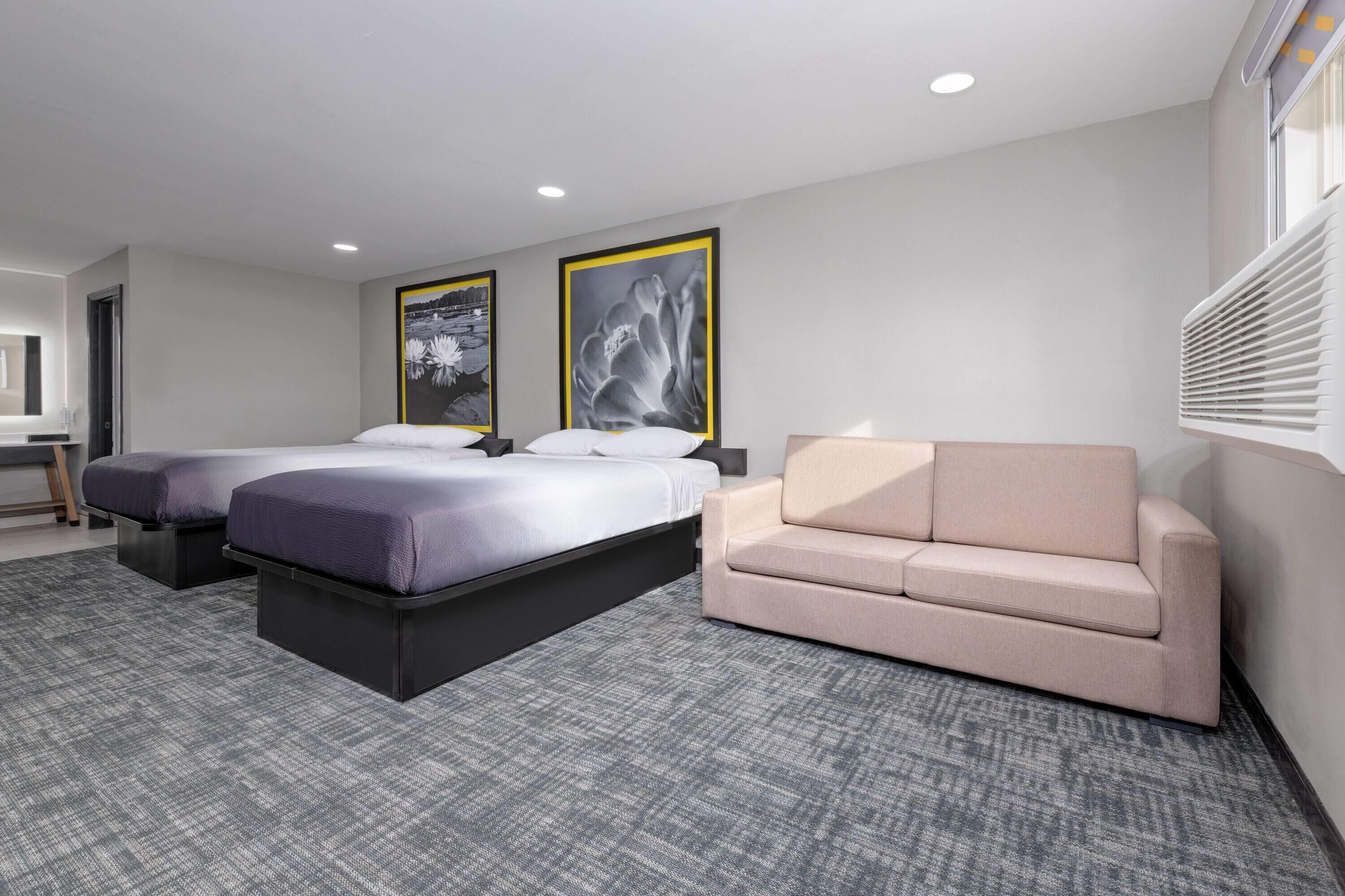 Une chambre d’hôtel avec deux lits, un canapé et une grande fenêtre équipée de stores.