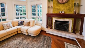 Un salon chaleureux avec un canapé d’angle beige, une cheminée et une grande fenêtre donnant sur un paysage verdoyant.