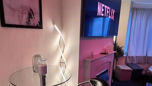 A modern living room with a glass table, black chairs, a flat-screen TV displaying 'NETFLIX', and a framed abstract painting.