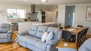 A modern living room with a grey sofa, a dining table with chairs, a kitchen area with appliances, and a large window. - Peppermint Rise - a luxurious retreat in Peppermint Grove Beach, WA. (Peppermint Grove Beach)