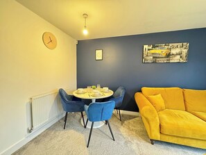 A dining area with a round table, blue chairs, a yellow sofa, a clock, and a framed picture on the wall.