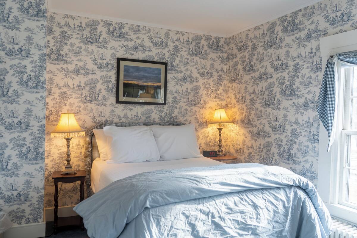 A bedroom with a bed, two bedside lamps, a clock, and a window with a checkered curtain.