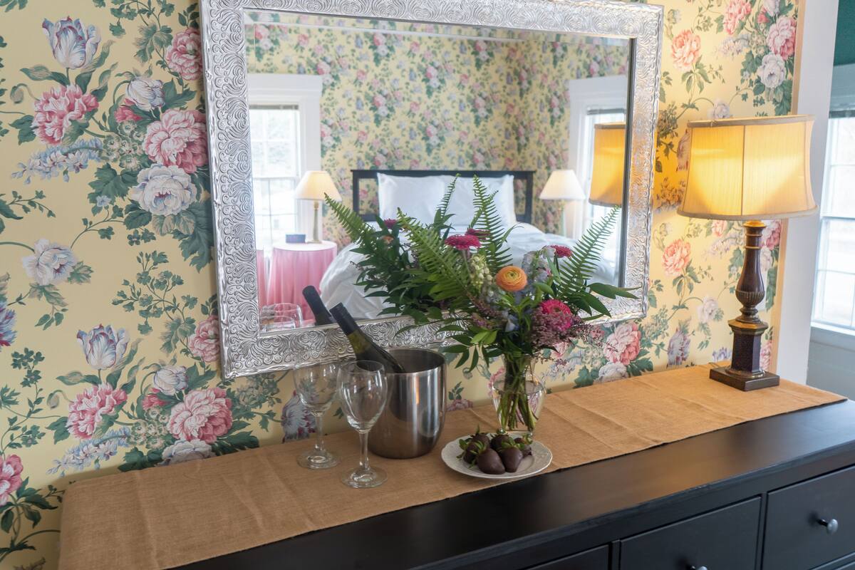 A room with a floral wallpaper, a bed with white bedding, a mirror, a lamp, a vase with flowers, and a bottle of champagne.