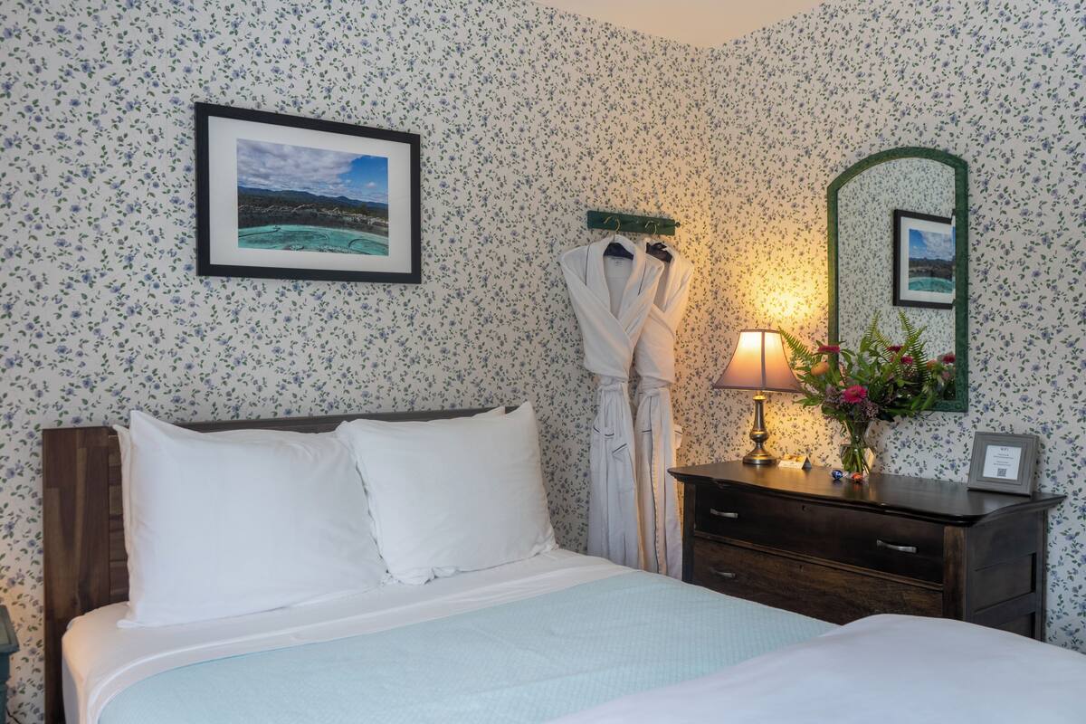 A neatly made bed with white bedding, a wooden headboard, a framed picture above the bed, a robe hanging on a hook, a bedside table with a lamp, and a mirror with a floral frame.