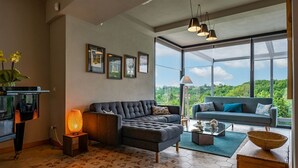 Ein modernes Wohnzimmer mit einer Couch, einem Couchtisch und einer Stehlampe. An den Wänden hängen gerahmte Bilder und ein großes Fenster bietet Blick auf Grünflächen.