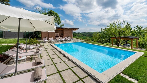 Ein rechteckiger Swimmingpool mit Liegestühlen und Sonnenschirmen, umgeben von einem Ziegelgebäude und viel Grün.
