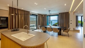 A modern kitchen with a central island, dining area with chairs, and large windows offering a view. - Villa Rora - Four Bedroom Villa, Sleeps 8 (Brela - Krug)