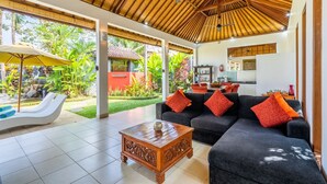 A spacious living area with a large sofa, a wooden coffee table, and a ceiling fan. There is a view of a lush garden and a red building outside.