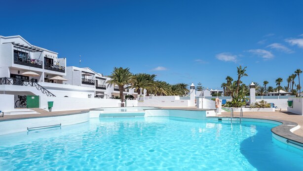 Una piscina con acqua cristallina e azzurra, circondata da edifici bianchi con balconi e palme.