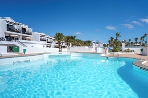 A swimming pool with clear blue water, surrounded by white buildings with balconies and palm trees. - 2 bedroom, 2 bathroom Apartment (Tías)